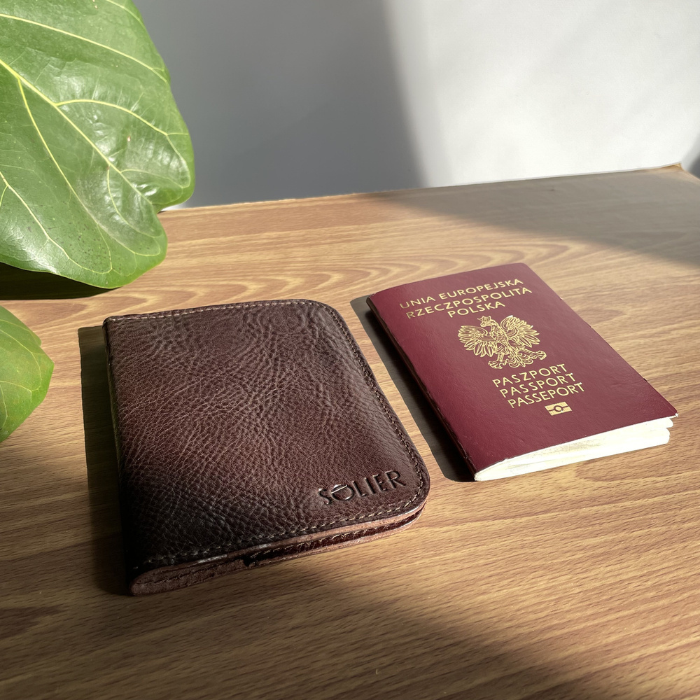 Brązowe etui na paszport Solier SW07A ze skóry naturalnej leżące obok paszportu na drewnianym blacie